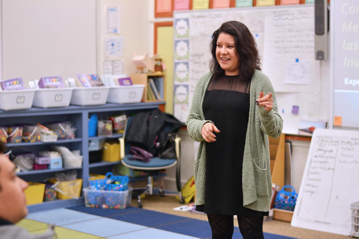 Woman in green cardigan and black shirt and pants giving lecture at front of K-12 classroom