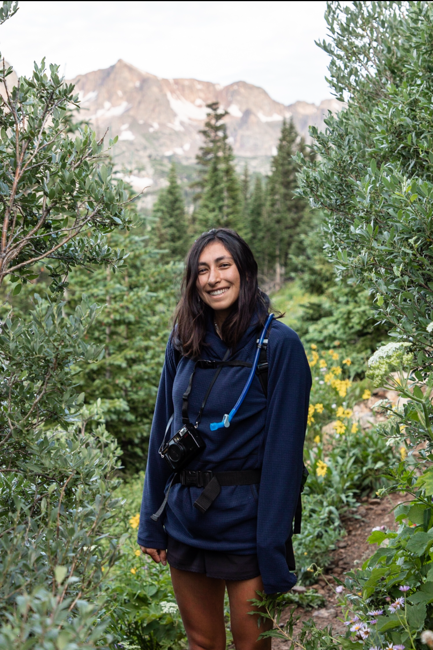 Woman with dark hair in dark clothing smiling and standing in front of woods and mountain view
