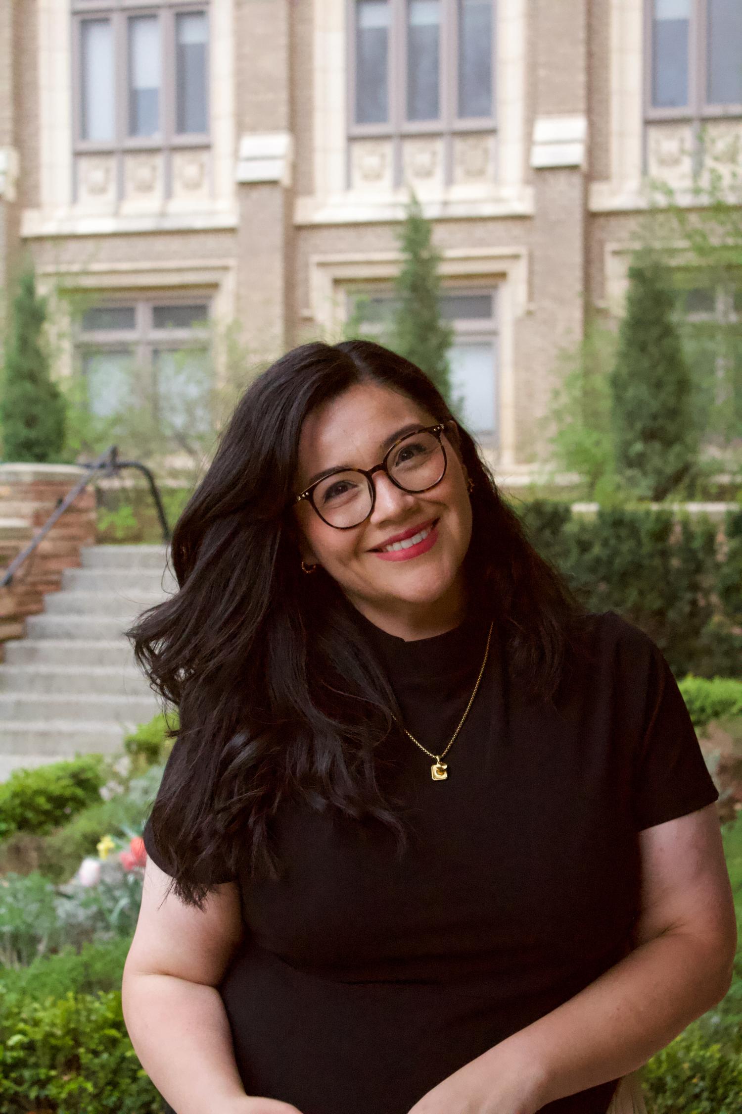 Celleny Juarez headshot; woman with glasses and long dark wavy hair in black shirt standing in front of campus building