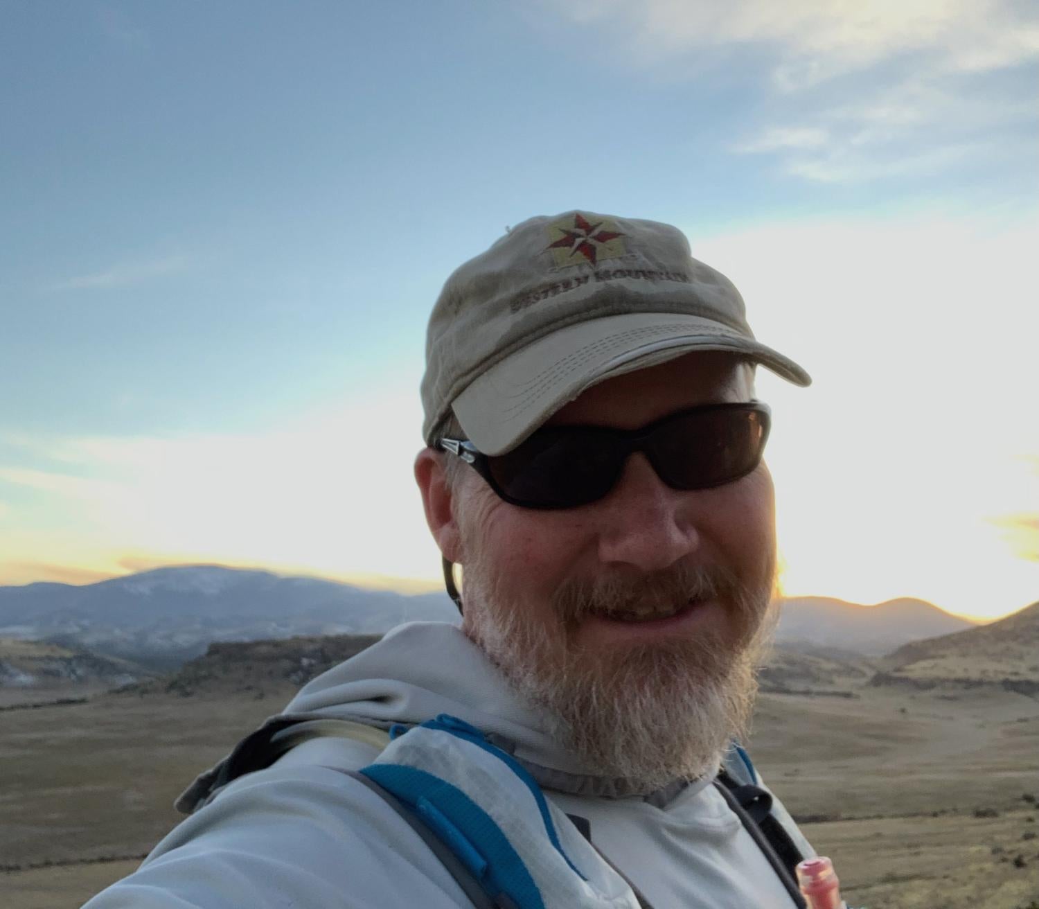 man in sunglasses and hat standing in front of mountain range shot as a selfie from shoulders up