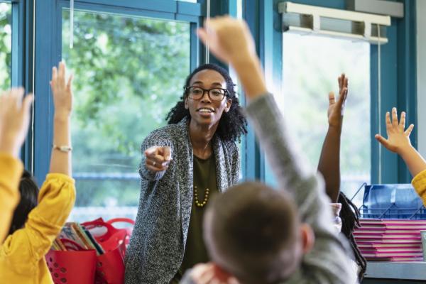 Woman with glasses at front of room pointing at students with raised hands at front of image