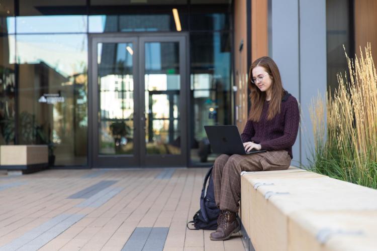 Image of a girl sitting outside an academic building working on a laptop on her lap.