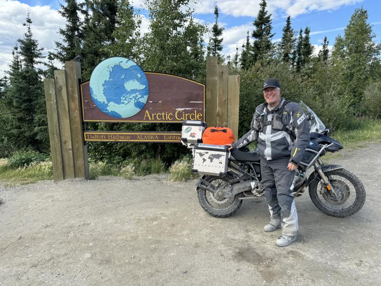 Gasta stands tall in front of a sign along the Dalton highway, noting his arrival at the edge of the Arctic Circle by motorbike with pride.  