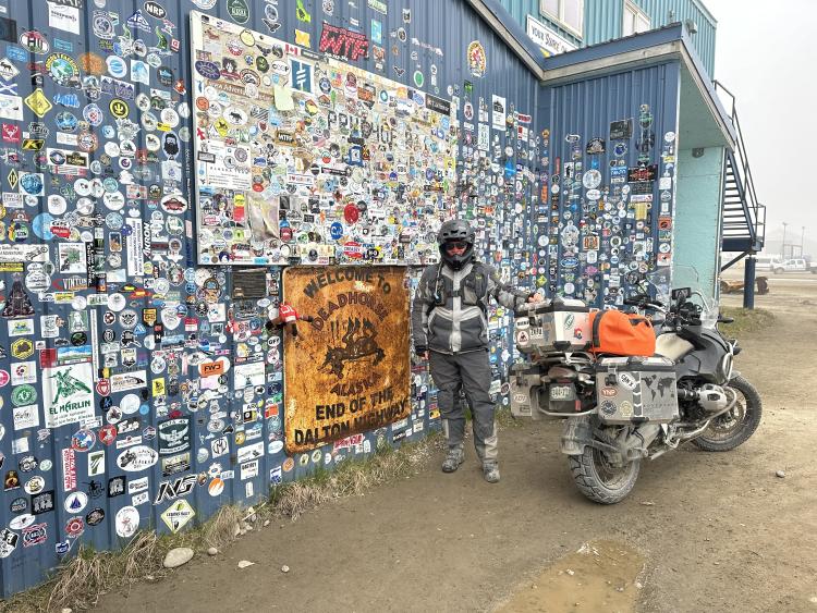 Gasta poses in full riding attire alongside his gear-laden motorcycle in front of an impressive international sticker wall in Deadhorse, AK.