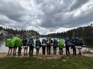 The group of thirteen participants stand with their backpacks on and back to the camera as they raise their trekking poles in excitement over a successful trip. They are positioned along the shore of a beautiful lake in the Sierra Nevada.