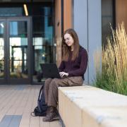 Image of a girl sitting outside an academic building working on a laptop on her lap.
