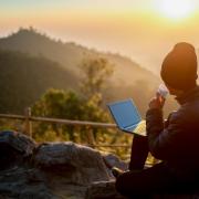 Man sipping from a mug outside while watching the sunrise over a mountain range. He has a laptop in his other hand.